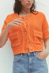 Chaqueta naranja de punto con cuello redondo y manga corta. Presenta bolsillos delanteros de parche con solapa y cierre frontal con botones. Detalle de pespuntes blancos a contraste.