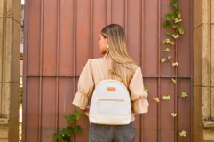 Mochila pequeña de cuero color gris claro y beige, con cierre superior y bolsillo frontal con cierre. Tiene asa de mano y correas ajustables para los hombros.