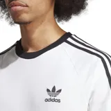 Remera blanca de corte clásico con cuello redondo y mangas cortas con ribete negro. Presenta tres rayas negras en los hombros y logo Adidas bordado en el pecho.