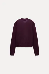 Chaqueta color borgoña de tejido de punto con lana y alpaca. Tiene cuello redondo con lazo de satén, manga larga y cierre frontal con botones. Presenta dos bolsillos de parche en el frente.