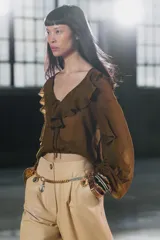 Blusa color marrón con escote en pico, manga larga abullonada con elástico en los puños y volantes en el escote y mangas.