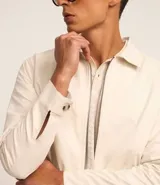 Campera color crema de sarga lisa, con cuello camisero, cierre frontal con cremallera, bolsillos delanteros, puños abotonados y bajo elástico.