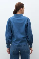 Camisa de jean de color azul medio, con cuello camisero, botones dorados y mangas largas abullonadas con puños ajustados.