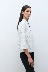 Blusa blanca con estampado de pequeños lunares negros, cuello camisero y botones frontales oscuros. Mangas tres cuartos con ligero vuelo en el puño.