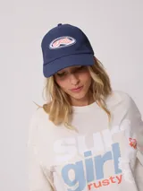 Gorra de lona blanca con visera azul marino y malla trasera azul marino. Presenta un logo bordado en el frente con letras rosadas y contorno azul marino.