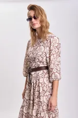 Vestido largo con estampado floral en tonos mostaza, marrón y blanco, con mangas largas abullonadas y falda con paneles. Cinturón marrón con tachas.