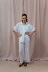 Camisa color crudo confeccionada en lino, de corte holgado, con cuello clásico, mangas cortas con dobladillo y cierre frontal con botones. Presenta detalle de pliegues verticales en la parte frontal.