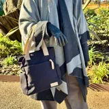 Cartera tipo tote de lona en color beige con correas de cinta marrón. Presenta un compartimento principal con cierre, un bolsillo frontal abierto y bolsillos laterales para mayor organización.