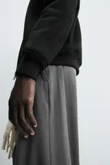 Pantalón gris de corte ancho con cintura elástica, pliegues frontales, bolsillos laterales y traseros de vivo. Incluye detalle de hebilla metálica en la trabilla y cierre frontal con cremallera y botón.