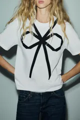 Camiseta blanca de corte recto, cuello redondo y mangas cortas. Presenta un lazo negro estampado en el frente.