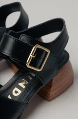 Sandalias negras de cuero con diseño minimalista y taco ancho geométrico en tono madera. La capellada lisa aporta una estética depurada y moderna, mientras que la pulsera con hebilla metálica ajustable asegura un calce firme y confortable.