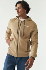 Campera de felpa color beige con capucha y cordón ajustable, cierre frontal, bolsillos con cierre, lettering estampado en la espalda y logo en grifa de goma.