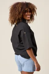 Camisa color beige claro, de corte clásico, con cuello, botones frontales, bolsillo en el pecho y mangas tres cuartos con presilla para sujetar.