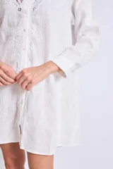 Vestido camisero corto color crudo, de lino, con cuello camisero, mangas largas y cierre frontal con botones. Presenta apliques de encaje en cuello y hombros.