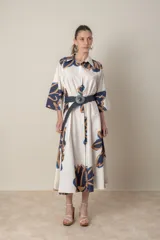 Vestido camisero midi de algodón color crudo con estampado de hojas abstractas en azul y ocre. Tiene cuello camisero, mangas tres cuartos y cinturón trenzado azul con hebilla metálica.
