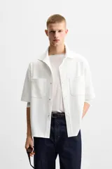 Sobrecamisa color beige de tejido elástico, con cuello solapa, manga corta y bolsillos de parche en el pecho. Cierre frontal con botones a presión.