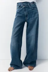 Pantalón de jean celeste de tiro alto, corte wide leg y largo hasta el tobillo. Presenta un diseño asimétrico en la cintura con cierre frontal cruzado con cremallera y botones metálicos.