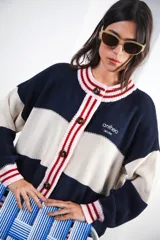 Cardigan de punto grueso con diseño a rayas horizontales en color crema, rojo y marrón. Presenta cuello redondo, cierre frontal con botones oscuros y ribetes a rayas marrones y celestes en cuello, puños y ruedo. Lleva un pequeño bordado con el logo de la marca en el pecho.