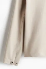 Campera color beige de tejido suave con cuello, cierre frontal, puños ajustables con botón a presión y bolsillos insertados al bies. Corte estándar y mangas forradas.