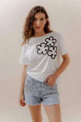 Remera blanca de algodón con cuello redondo y manga corta. Presenta un aplique frontal con tres flores estilizadas bordadas en hilo negro.
