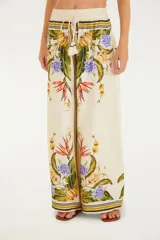 Pantalón de viscosa con estampado tropical de flores y hojas en tonos naranja, amarillo, verde y violeta sobre fondo blanco. Tiene cintura alta con elástico y cordón ajustable con borlas.