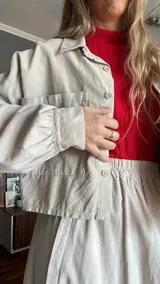 Camisa corta color beige de pana con cuello camisero, cierre frontal con botones, manga larga con puños elásticos, bolsillo de parche en el pecho y corte a la cintura con elástico.