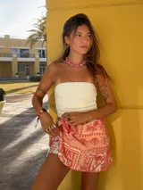 Falda pareo corta, color coral, con estampado de temática playera que incluye sellos postales, conchas marinas y texto. Se anuda en la cintura.