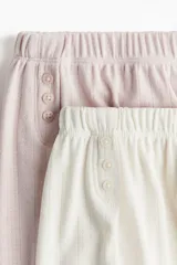 Pack de dos shorts de pijama en punto pointelle: uno rosa y otro blanco. Ambos tienen corte holgado, elástico en la cintura, cierre decorativo con botones y bajo sobrehilado.