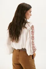 Camisa blanca de manga larga con bordados marrones en hombros y mangas, cuello en V y cierre frontal con botones.