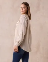 Blusa color beige de lino y rayón, con cuello a la base, escote en V y abertura con botones. Tiene bolsillos en la parte delantera y mangas largas regulables con presilla y botón.