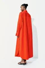 Vestido camisero largo color naranja, de corte amplio, con cuello clásico, cierre frontal con botones, mangas largas con puños abotonados, bolsillos laterales y tajos laterales pronunciados.
