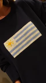 Sweater de tejido de punto color gris oscuro, con cuello redondo y mangas largas holgadas. Presenta un parche tejido en el pecho con la bandera de Uruguay.