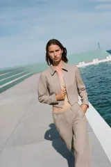 Chaqueta corta color beige con cuello solapa, manga larga y cierre frontal con cremallera.