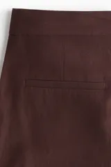 Pantalón sastrero color marrón chocolate, tobillero, de tiro alto, con cierre y botón. Presenta bolsillos al bies, bolsillos traseros decorativos y piernas amplias con pliegues adelante y atrás.