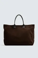 Bolso tipo shopper marrón oscuro, fabricado en cuero con acabado serraje. Tiene un compartimento principal con cierre magnético y un bolsillo interior con cremallera. Cuenta con dos asas de mano de cuero.