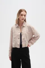 Chaqueta corta de estilo camisa, color beige claro con finas rayas verticales blancas. Presenta cuello camisero, manga larga y cierre frontal con botones de color claro. Tiene dos bolsillos de parche con solapa en el pecho.