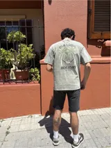 Remera gris de corte amplio con estampa en la espalda de palmeras, ángel en una hamaca y texto 'Paradise Holiday'.
