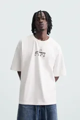 Camiseta blanca de corte holgado, cuello redondo y manga corta. Estampado frontal y trasero con gráficos y textos combinados en color negro.