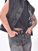 Chaleco de denim azul lavado, sin mangas, con cuello camisero, botones frontales y bolsillos de parche en el pecho.