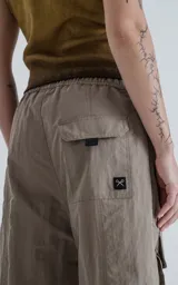 Bermuda técnica color arcilla (marrón claro/beige) de calce oversize, confeccionada en poliéster liviano. Presenta múltiples bolsillos cargo con tapa y un cinturón de ajuste con hebilla plástica negra. Incluye detalles reflectivos.