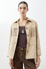 Chaqueta color beige de simil gamuza con flecos en mangas y ruedo, con cuello camisero y cierre frontal con botones.