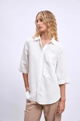 Camisa blanca de manga tres cuartos, con cuello camisero y corte holgado. Presenta un bolsillo de parche en el pecho izquierdo.