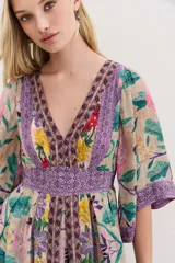 Vestido largo de viscosa con mangas al codo y falda amplia. Presenta un estampado floral vibrante sobre fondo beige, con detalles y bordes en tonos morados y patrones geométricos. Tiene escote en V y cierre trasero.