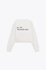 Sudadera corta color crudo de algodón, con cuello redondo, mangas largas y texto estampado en el frente que dice "IN MY YEEHAW ERA".