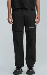 Pantalón cargo negro de corte recto y holgado, con bolsillos grandes con solapa en los laterales de las piernas y un pequeño parche blanco con texto en uno de ellos.