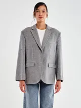Blazer de mezcla de lana color gris, con solapas de muesca y bolsillos de parche delanteros. La prenda está adornada con apliques tipo joya plateados distribuidos por toda la superficie.