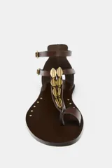 Sandalia plana de cuero color marrón con tira delantera con adorno metálico dorado y doble tira con hebilla al tobillo.