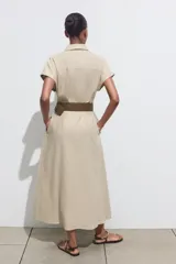 Vestido midi camisero color beige, confeccionado en lino. Presenta cuello solapa, mangas cortas con vuelta, jaretas delanteras, bolsillos laterales ocultos en costura, cierre frontal con botones y cinturón trenzado marrón.