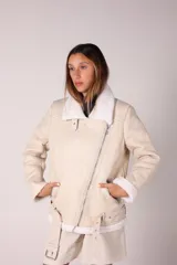 Campera gamulán color blanco hueso con interior de corderito. Tiene corte oversized, cierre cruzado metálico, bolsillos laterales con cierre, cinturón con hebilla ajustable y dobladillo y puños con borde de corderito a la vista.