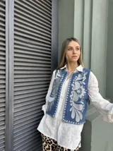 Chaleco de jean azul con bordado floral blanco y ribete de tachas plateadas.